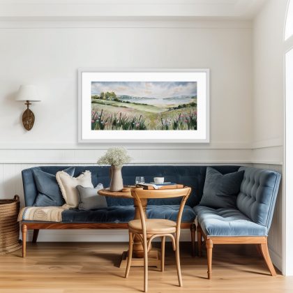 Watercolour landscape painting shown in a living room setting with blue chairs. The painting shows a wide lake and headlands from a hilly meadow. In the immediate foreground, detailed wild grasses, pink poppies, and pale blue cornflowers grow along a rusty barbed wire fence with dark posts. Beyond the fence, rolling, soft green and ochre fields with scattered trees lead down to a large body of calm water. A small white cottage sits near a cluster of green trees on the far left. On the distant shore, rolling hills covered in a soft wash of green foliage outline the horizon. The sky is filled with swirling, pale grey, blue, and faint apricot-orange clouds. The entire scene has the soft, blended edges and fluid quality typical of watercolour painting.