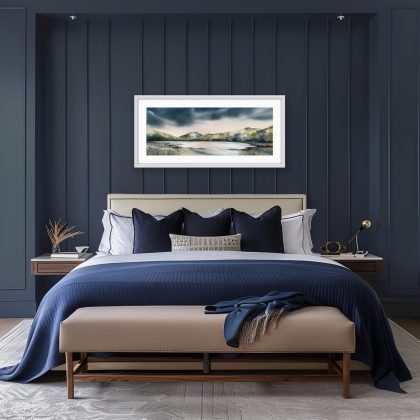 A horizontal watercolour painting of a tranquil lake surrounded by rolling green and earthy-toned hills under a dramatic, dark storm sky shown in a bedroom setting with deep blue walls and bed linen. Thick, moody indigo and charcoal clouds dominate the top half of the composition. In the centre, the calm water reflects a bright, pale light, with two small sailboat masts visible in the distance. The foreground features textured shoreline grasses and dried seed heads in muted tones of tan and grey. The style is atmospheric with soft, wet-on-wet watercolour washes creating a sense of mist and depth.