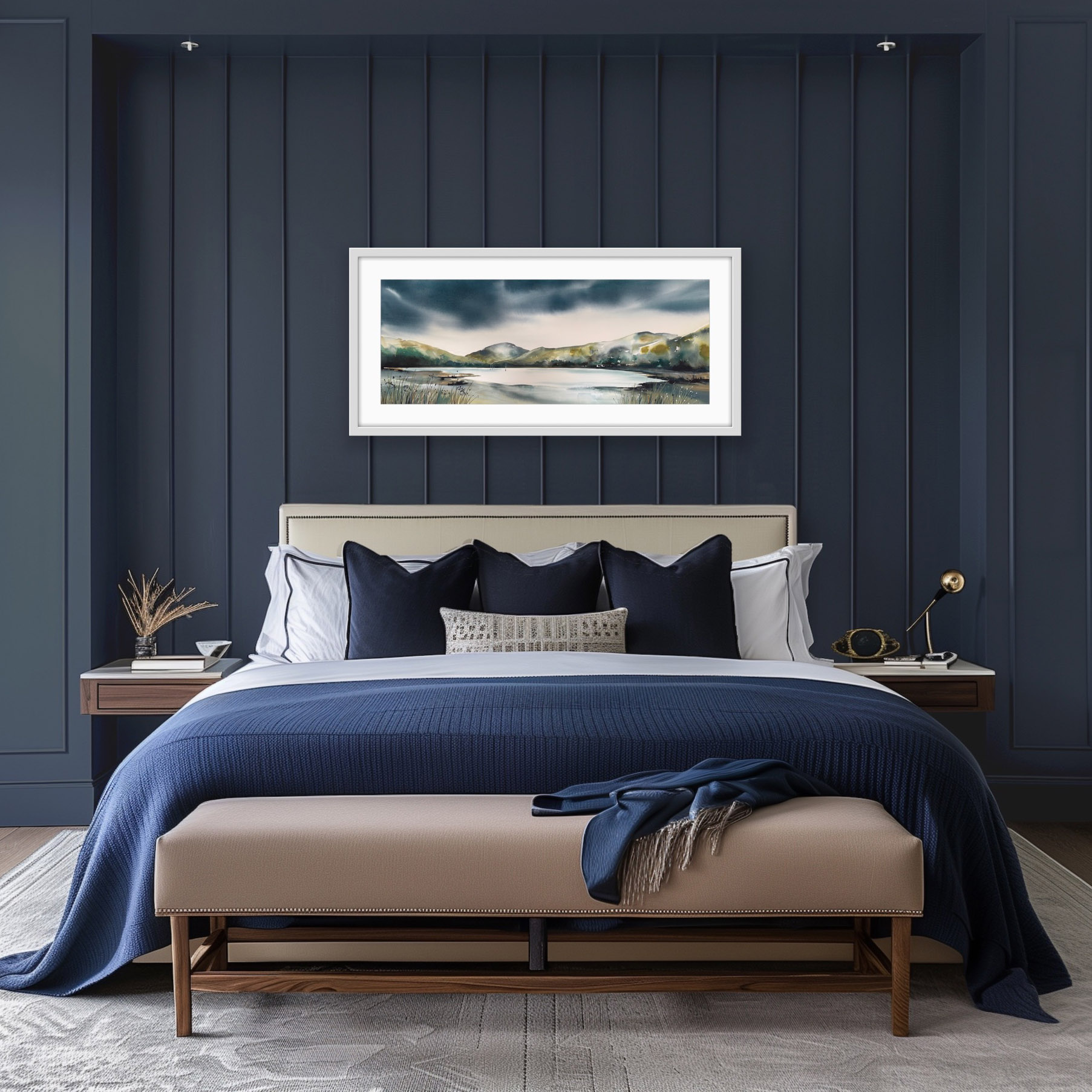 A horizontal watercolour painting of a tranquil lake surrounded by rolling green and earthy-toned hills under a dramatic, dark storm sky shown in a bedroom setting with deep blue walls and bed linen. Thick, moody indigo and charcoal clouds dominate the top half of the composition. In the centre, the calm water reflects a bright, pale light, with two small sailboat masts visible in the distance. The foreground features textured shoreline grasses and dried seed heads in muted tones of tan and grey. The style is atmospheric with soft, wet-on-wet watercolour washes creating a sense of mist and depth.