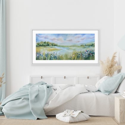 A horizontal watercolor landscape painting of a lush green meadow and river under a soft blue and cloudy sky. In the foreground, detailed wildflowers including blue cornflowers and white daisies grow among tall blades of grass. The background features misty green hills and a cluster of trees in soft, blurred tones. The painting is in a bedroom setting with white walls and pale blue bedding.